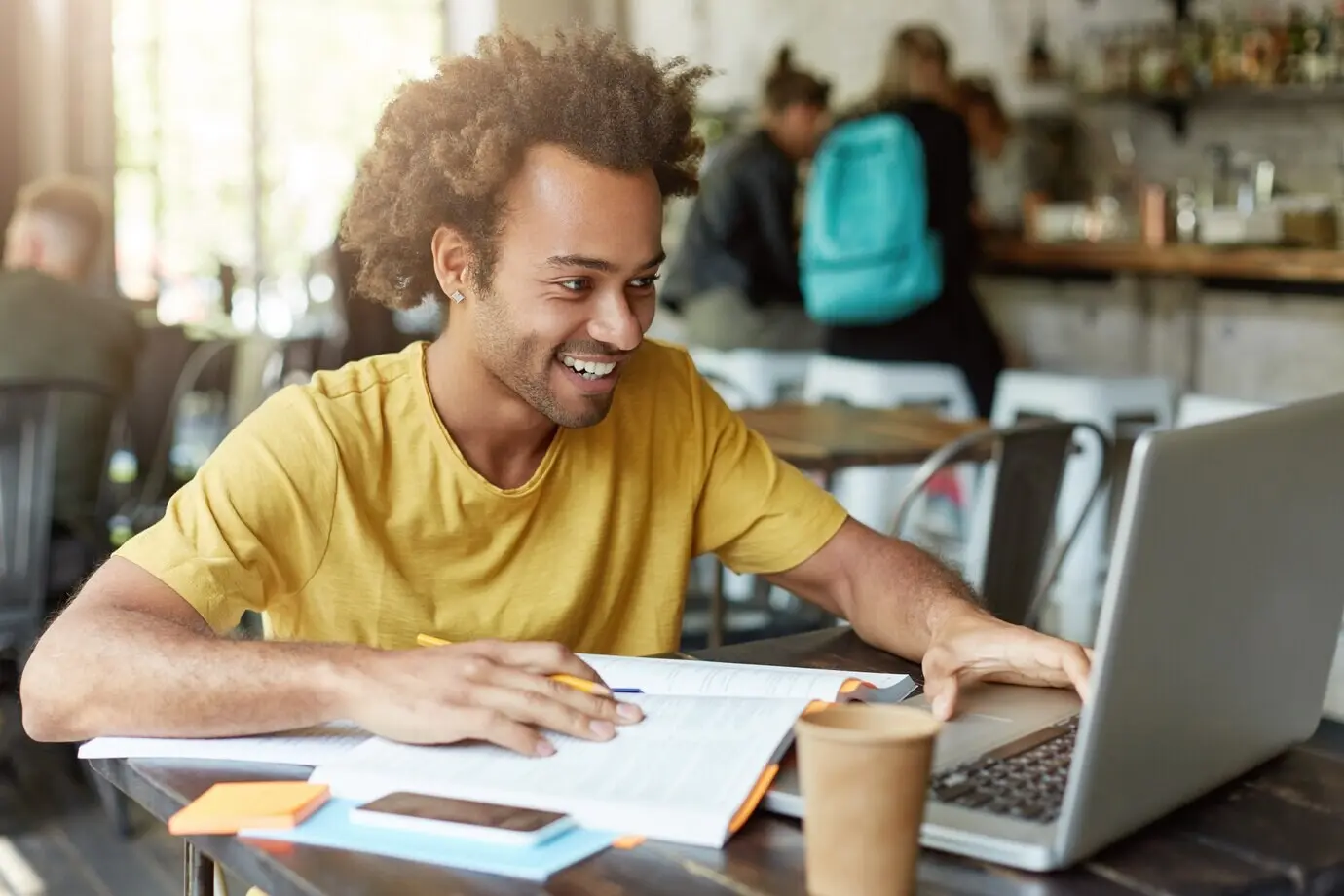 Innenaufnahme eines fröhlichen männlichen Studenten mit lockigem Haar, leger gekleidet, der in einer Cafeteria sitzt, beim Lernen mit modernen Technologien arbeitet, lächelnd in sein Notebook blickt und eine Nachricht von einem Freund erhält.